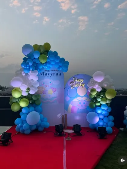 An outdoor rooftop birthday party setup for Mayyraa. The decoration features two arched backdrops with a Sanrio character theme and blue and green balloon arrangements.