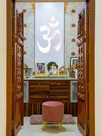 This pooja room features beautifully carved traditional wooden doors that open to reveal a modern, serene space with a backlit Om symbol, blending classic craftsmanship with contemporary design.