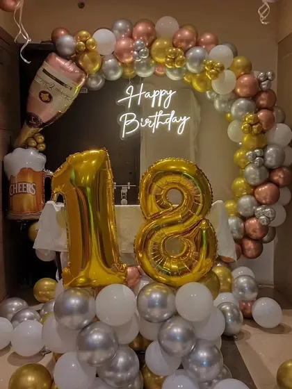 A festive 18th birthday decoration with a balloon arch in gold, silver, and white. The theme is enhanced with champagne bottle and beer mug foil balloons.