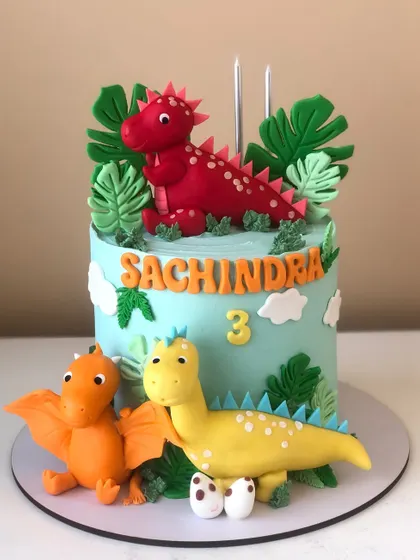 A colorful dinosaur cake for a third birthday, with three handcrafted dinosaur figures in a prehistoric setting.