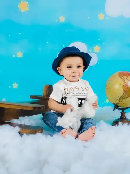 A baby boy in a hat sits with a toy airplane, surrounded by clouds, dreaming of his next big trip.