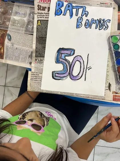 A student paints a colorful sign for her "Bath Bombs" stall. The flea market project allowed children to take ownership of their work, from product creation to marketing.