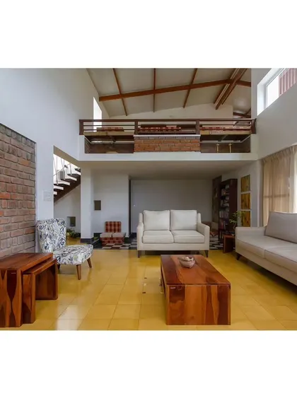 The living room in 'Malenadu Mane' features a double-height ceiling with an overlooking mezzanine. The use of exposed brick and natural wood furniture gives the space a warm, rustic, and inviting feel.