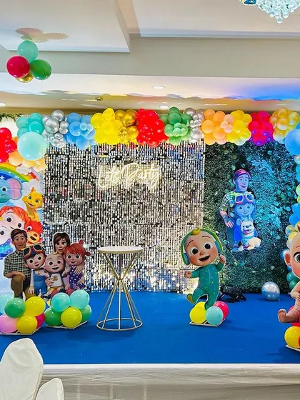 A Cocomelon-themed stage setup with a silver shimmer wall and a blue carpet. The decor includes character cutouts and a light-up Ferris wheel.
