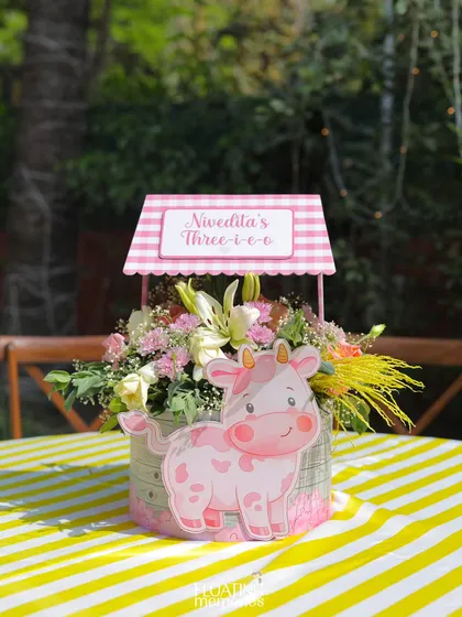 Our custom centerpieces featured fresh flowers in a basket, adorned with a pink gingham wishing well and a cute cow cutout. This detail carried the 'Three-i-e-o' farm theme to every table.