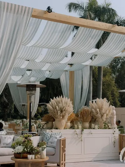 A lounge area defined by a wooden pergola with a striped fabric ceiling. The neutral-toned furniture is accented with printed cushions, creating a relaxed and sophisticated space.