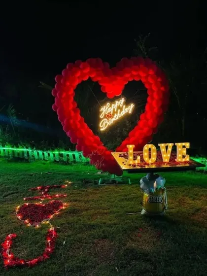 A grand romantic gesture in a garden at night. A large heart-shaped balloon arch surrounds a 'Happy Birthday' neon sign, with 'LOVE' marquee letters and a path of rose petals.