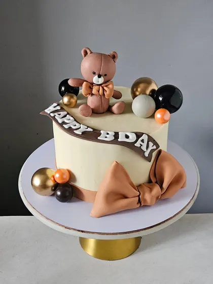 A stylish and sweet teddy bear cake with a modern, earthy color palette. The design features a "Happy Bday" banner, a large fondant bow, and decorative spheres in black, gold, and brown.
