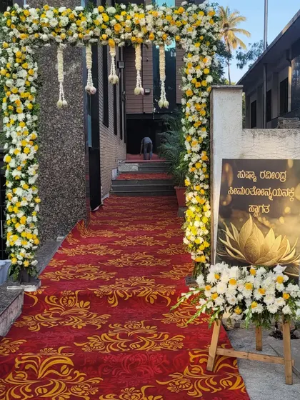 I create welcoming entrances that set the tone for the event. This one features a grand floral arch of white and yellow flowers over a red carpet, with a custom welcome sign in Kannada to greet the guests.