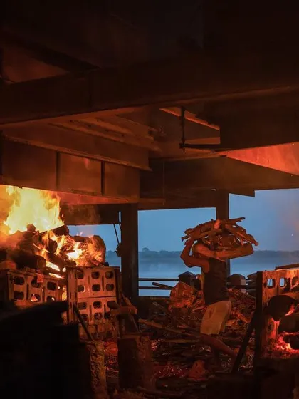 Two funeral pyres burn side-by-side inside the electric crematorium structure at Manikarnika, with the Ganges visible in the background. This shot documents the more modern facilities alongside traditional ones.