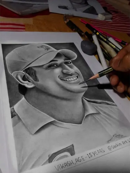 A photo of my hand adding the final touches to the MS Dhoni portrait, showing the tools I use and the drawing on my desk.