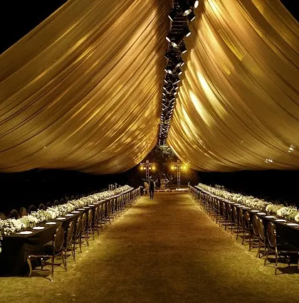 A dramatic sit-down dinner setup with long tables, black linens, and a striking gold fabric canopy. The balance between dark elegance and warm light creates a truly memorable ambiance.