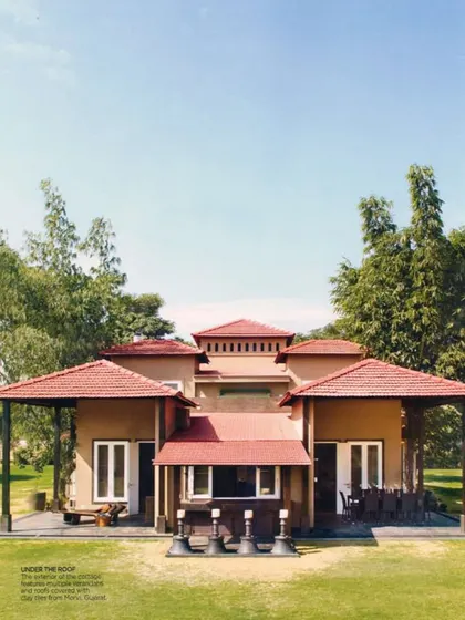 An exterior shot of the Aishbagh cottage featured in a magazine, highlighting its distinctive red-tiled roof and symmetrical architecture. The home is designed to sit comfortably within its lush, green surroundings.
