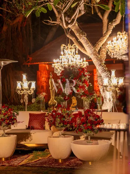 A cozy lounge nook under a tree, adorned with chandeliers. The white sofas are accented with red cushions and surrounded by arrangements of red roses, creating a romantic seating area.