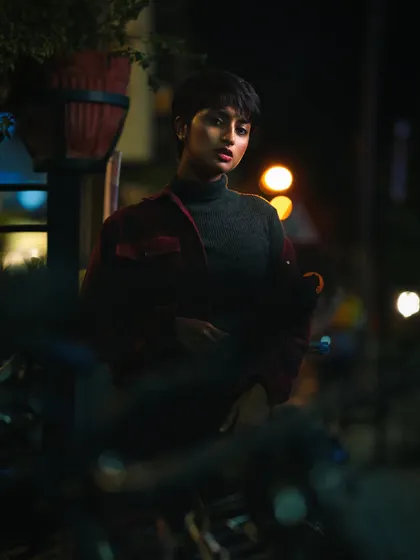 A captivating night portrait of a woman with short hair, surrounded by the soft, blurry lights of the city. The dark, moody tones and her direct gaze make for a powerful and atmospheric shot.