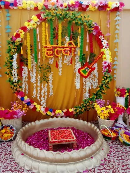A festive Haldi setup featuring a circular floral frame, a guitar prop, and a traditional 'urli' filled with rose petals.