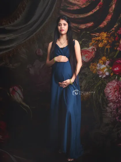 A full-length portrait in a simple blue gown against a classic painted floral backdrop.