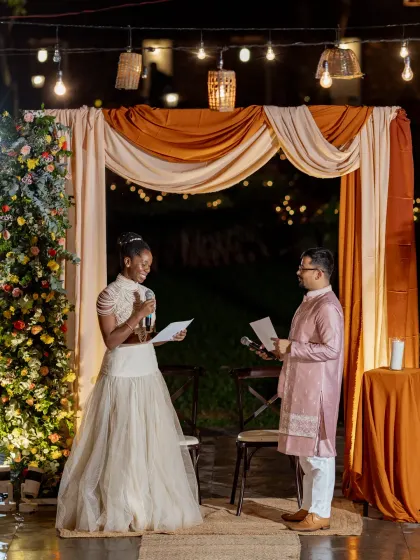 The couple exchanges their vows in a heartfelt, intimate ceremony, with a simple yet elegant backdrop of drapes and string lights.