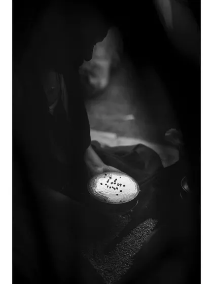 A black and white photo focusing on a bowl of offerings held by a devotee. The framing is intentionally tight and obscured, creating a mysterious and intimate feeling that centers on the act of giving.