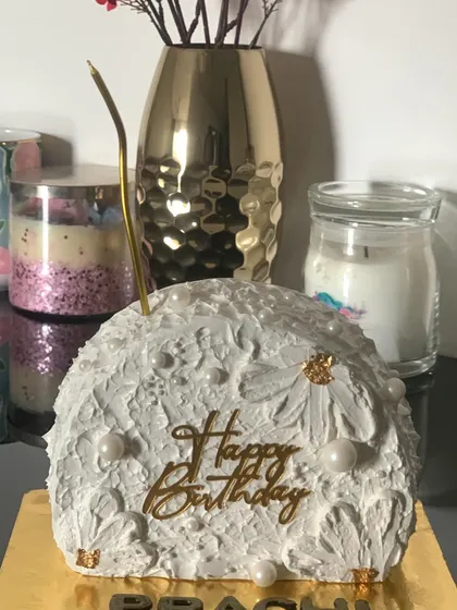 A modern "top forward" style cake. The unique half-dome shape is decorated with a textured white icing, pearls, and a gold birthday topper.