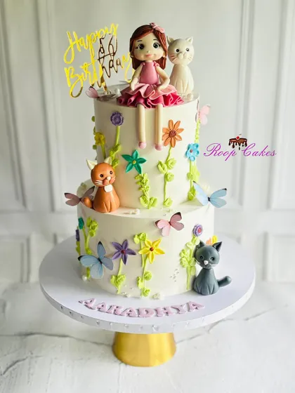 Whiskers, flowers, and birthday magic. This adorable two-tier cake is bursting with charm, featuring playful cats, blooming flowers, butterflies, and a sweet little doll topper.