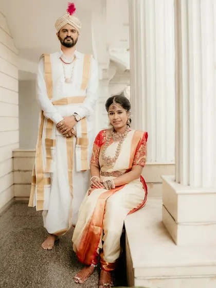 A regal portrait of the bride and groom in their traditional South Indian wedding attire. The groom stands tall while the bride is seated, creating a classic and elegant composition.