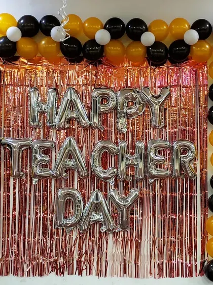 A decoration for a Teacher's Day celebration. This backdrop features a rose gold shimmer curtain, "Happy Teacher Day" foil letters, and a frame of black, gold, and white balloons.