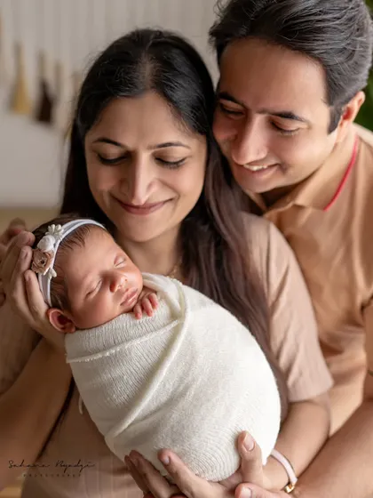 A close-up portrait of new parents holding their swaddled baby. Their expressions of love and awe are the central focus of this intimate and emotional photograph.