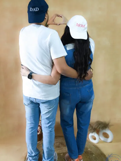 A fun and creative shot of a couple from behind, wearing "Mom" and "Dad" caps and making a heart shape with their hands.