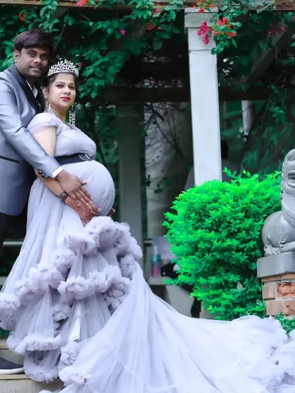 A loving couple's maternity portrait in a garden setting, with the woman in a grey ruffled gown and the man in a matching silver suit.