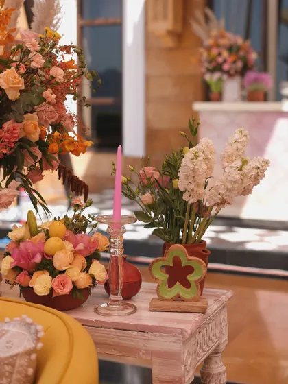 A close-up of a side table with a pink candle, a flower-shaped cookie, and a beautiful arrangement of pink and orange flowers.