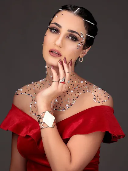 A high-fashion editorial look combining a bold red dress with intricate glitter and pearl body art. The focus is on creating a luxurious and artistic beauty statement.