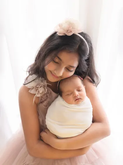 A tender moment between an older sister and her newborn sibling. The soft, high-key lighting and gentle embrace create a portrait that is pure, simple, and full of love.