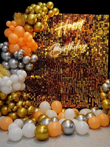 A close-up of a shimmer wall backdrop, a popular choice for creating a glamorous photo booth. This one uses a gold sequin panel with a 'Happy Birthday' neon sign, complemented by a balloon garland in orange, silver, and gold.