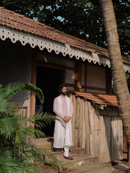 The groom stands outside a rustic, traditional Goan house, his simple yet elegant attire fitting perfectly with the charming, earthy setting.