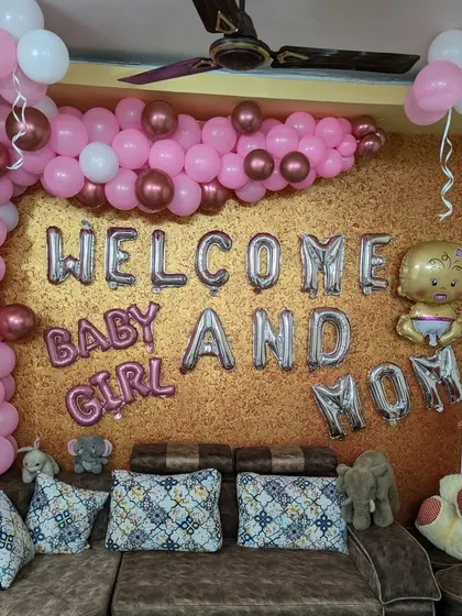 This welcome home decoration for a baby girl and mom includes a partial balloon arch and foil letters, complemented by soft toys on the sofa.