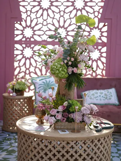 A cozy lounge area with pink velvet chairs, carved wooden tables, and soft floral arrangements against a geometric-patterned screen.