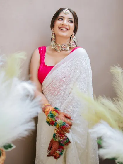 A beautiful portrait of the bride in her unconventional Haldi attire, a white saree paired with a bright pink blouse, surrounded by pampas grass decor.