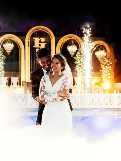 A spectacular poolside reception entrance. The couple is surrounded by low-lying fog and cold sparklers, creating a dreamy and dramatic effect against the elegant arched backdrop.