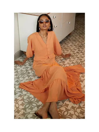 A high-angle shot of the model seated on a beautifully tiled floor. The sunglasses and relaxed pose give this image a chic, vacation-ready vibe, perfectly showcasing the flowing peach dress.