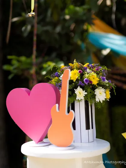 A vibrant centerpiece from a music-themed party, featuring a colorful floral arrangement in a piano-key vase, flanked by a bright pink heart and an orange guitar cutout.