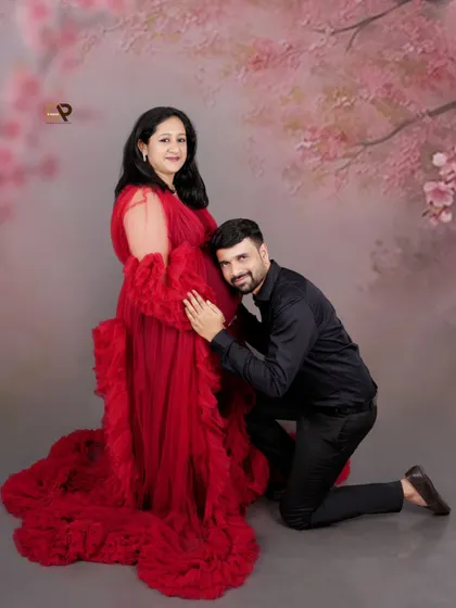 A sweet moment between the expecting parents. The dad-to-be listens to the baby bump, while the mom-to-be glows in a vibrant red gown against a soft, cherry blossom-themed backdrop.