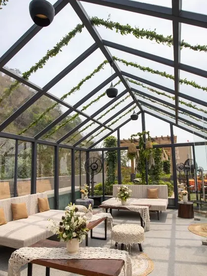 A beautiful glasshouse seating area, decorated with white sofas, macrame-topped stools, and natural-textured runners. The space is filled with light and greenery, creating a serene and rustic feel.