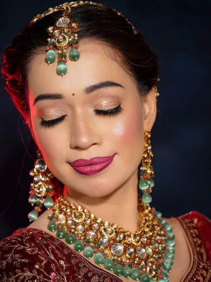 A retro-inspired bridal look with a modern twist. The soft, shimmery eyes and bold magenta lip create a stunning contrast with the deep red velvet lehenga and green jewelry.