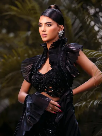 A striking outdoor shot featuring a structured, all-black ensemble with dramatic ruffled shoulders. The natural greenery provides a beautiful contrast to the high-fashion look.