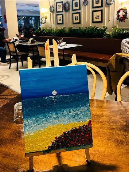 A finished knife painting of a beach scene, resting on an easel in a cozy cafe setting, waiting to be taken home.