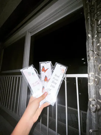 Holding up a set of custom bookmarks against a night sky. These feature pretty butterfly designs and quotes like "Read more, sleep less."