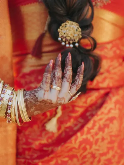 A close-up of the bride's hand, adorned with intricate henna and traditional bangles, resting against her vibrant red saree.