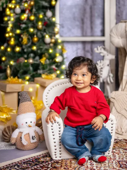 A happy baby boy in a bright red sweater, sitting contentedly in his chair amidst the festive Christmas decorations.