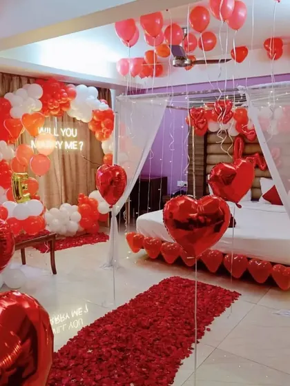 A grand 'Will You Marry Me?' proposal setup in a room, with a red rose petal path, a neon sign, and a heart-shaped balloon arch.
